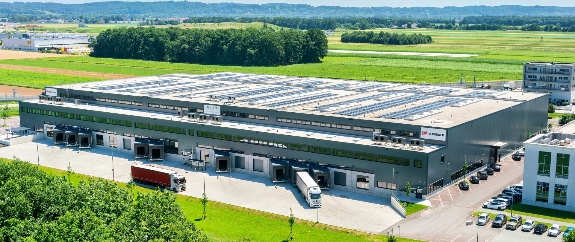 Luftaufnahme eines modernen Logistik- und Gewerbeparks mit Photovoltaikanlage am Dach und Lkw-Anlieferzonen – Immobilie der Pletzer Gruppe mit DB Schenker als Nutzer, eingebettet in grüne Umgebung.